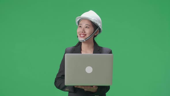Smiling Asian Female Engineer Looking Around While Using Laptop In The Green Screen Studio alt