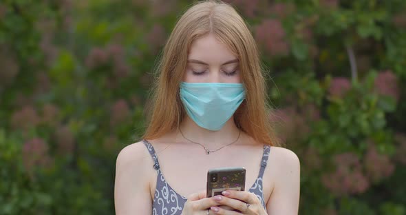 A young girl in a protective medical mask looks at the phone and sends messages