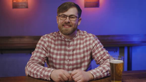 Portrait of a Happy Young Man in Glasses in Pub with Glass of Beer in Hand at Bar Counter alt