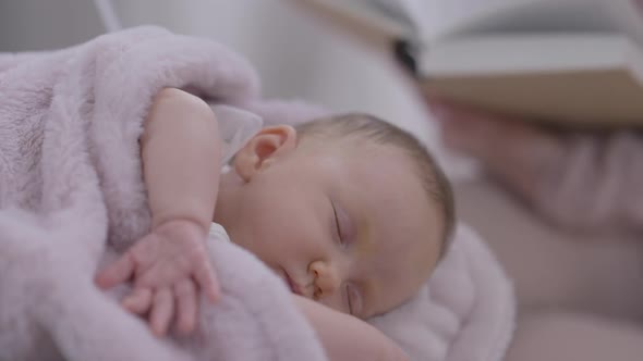 Closeup Face of Sleeping Infant with Blurred Woman Turning Book Pages at Background alt
