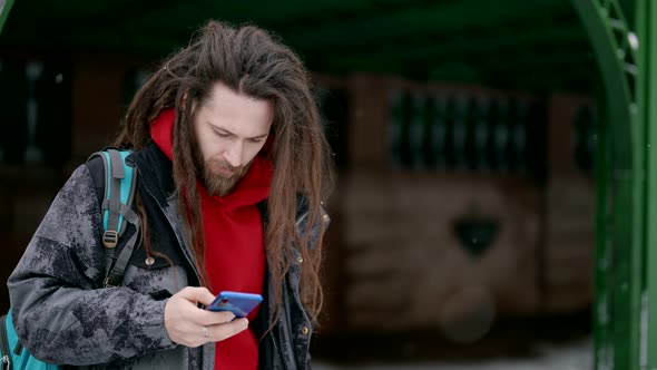 Trendy Man with Dreadlocks is Using Smartphone on Street in Winter Swiping and Scrolling Display alt