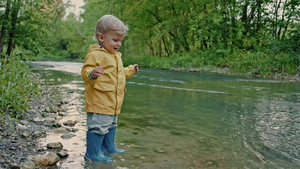 Cute Baby in Raincoat and Rubber Boots Has Fun By River, Throws Pebbles and Laughs, Funny Boy Learns alt