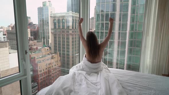Girl Wrapped in Blanket in Bedroom Sits on Bed Near Window View at New York
