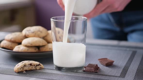 Woman Pours Milk Into Glass with Fresh Made Homemade Soft Chocolate Chip Cookies on Background alt
