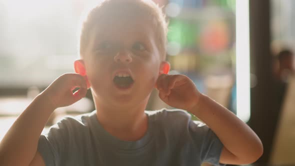 Little Boy with Open Mouth Closes Ears By Fingers in Room alt