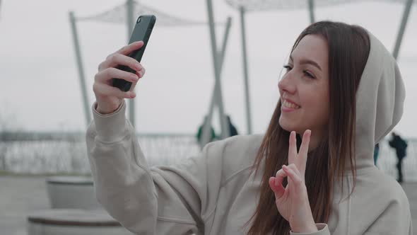 Close Up View of Cheerful Young Pretty Woman in Beige Hoodie Taking Selfie on Smartphone and alt