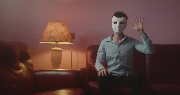 Anonymous man with mask greets with his hand while sitting on the red sofa at home alt
