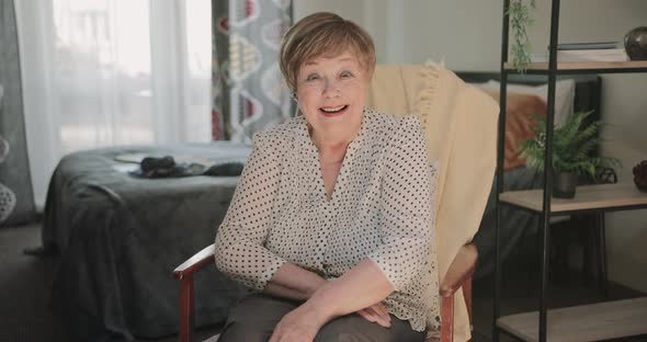 Front View of Cheerful Woman in 60s Sitting Comfortable at Chair and Possing To Camera alt