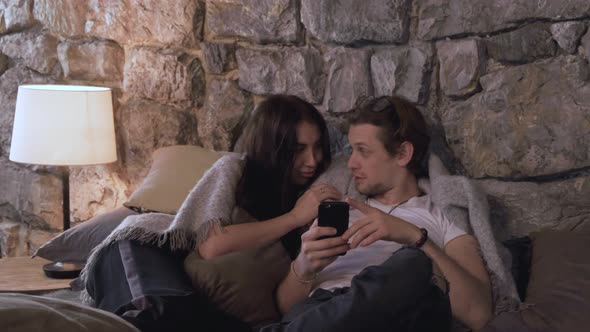 A Young Couple Are Lying In Bed Chatting Looking At Photos On Their Smartphone alt