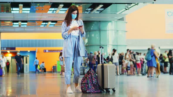 Woman in Mask, Standing at Airport, with Luggage, Waiting To Board the Plane. She Is Checking Flight alt