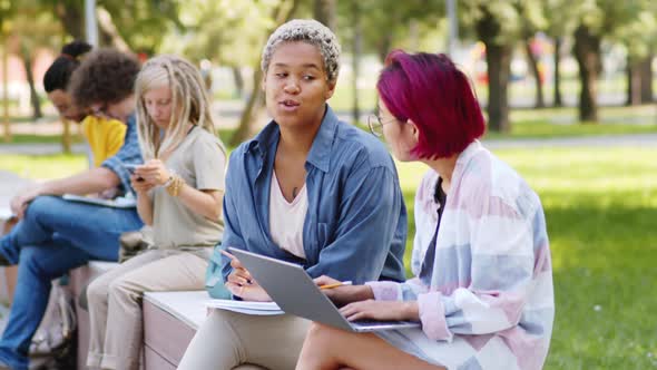 Two Multiethnic Female Students Discussing Project Outdoors in Park alt