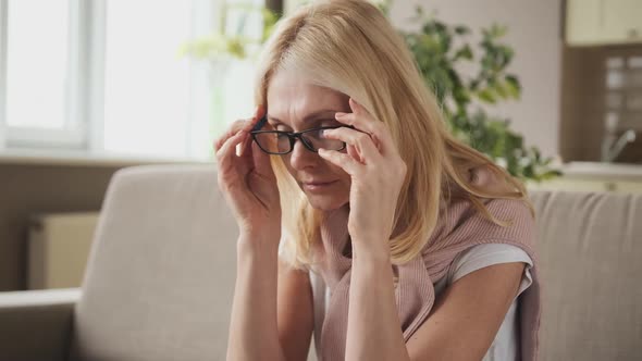 Middleaged Woman Puts on Glasses with Her Hands Sitting on the Couch at Home alt