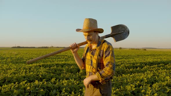 Farmer Man Holding a Shovel in His Hand Walking Across the Green Soybean Field. Eco Smart alt