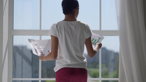 Back View of Young Slim Woman Walking with Business Graphs to Window Throwing Away Paperwork Looking alt
