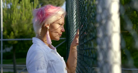 Bold and Blonde Woman Is Looking Through Metal Fence Grid of Open Sporty Area alt