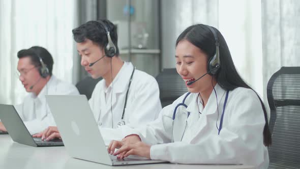 Three Asian Doctors With Stethoscopes In Headsets Working As Call Centre Agents While Speaking alt