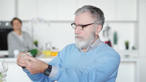 Portrait of Senior Man in Eyeglasses Examining Dollar Banknote Indoors alt