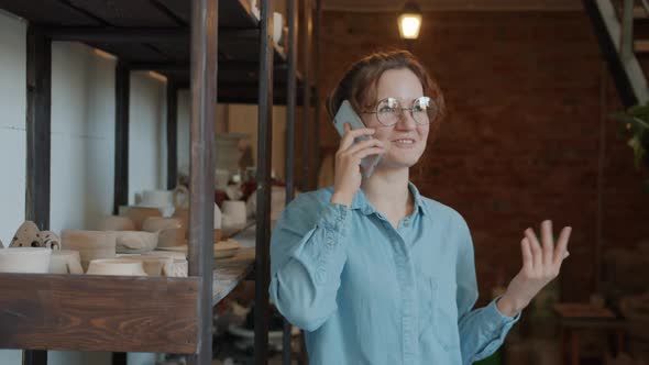 Happy Young Lady Speaking on Mobile Phone in Pottery Workshop Smiling alt