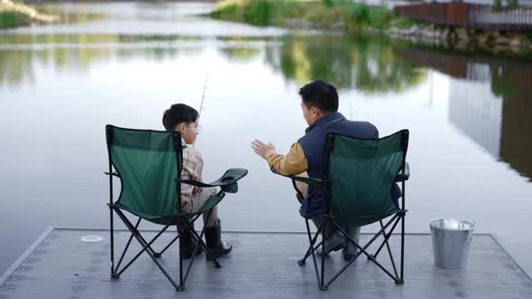 Dad and Son Sit in Fishing Chairs on the Pier on the City Lake and Father Tells Funny Story the Son alt