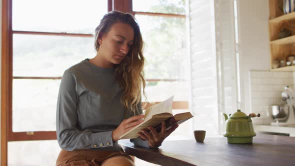 Happy caucasian woman sitting in cottage kitchen reading book, with teapot on counter alt