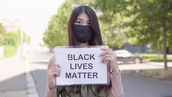 Young Asian Woman Face Mask Shows Black Lives Matter Sign To Camera Park Trees alt