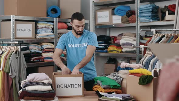 Caucasian Bearded Male Volunteer Sorting Clothes and Putting It Into the Cupboard Box at the alt