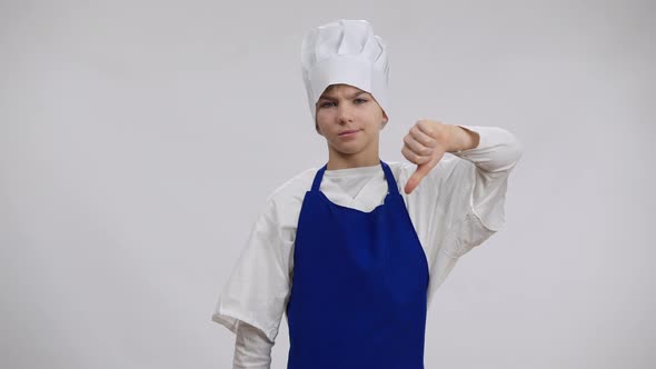 Caucasian Boy in Blue Apron and White Chef Hat Posing at Background Showing Thumb Down alt