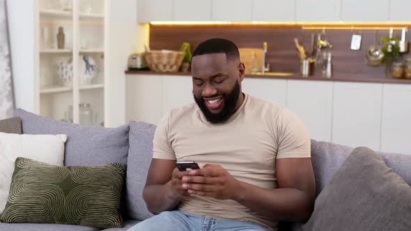 Handsome Positive African American Man in Casual Wear Sits at Home on Sofa Using His Smartphone alt