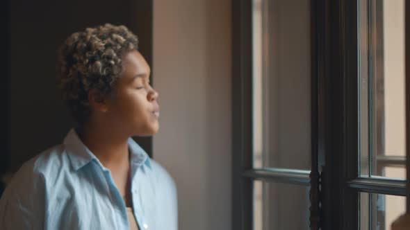 Young African Woman Standing By Window at Home Looking Happy and Content alt