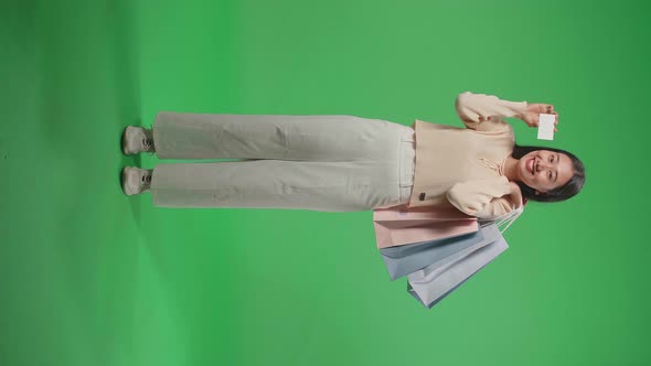 Full View Of Asian Woman Holding A White Card And Shopping Bags Standing In Front Of Green Screen alt