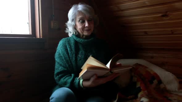 Elderly Woman Sits in a Wooden Room and Flatters a Book