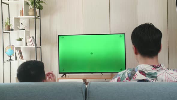 Father And His Son Are Watching Green Mock-Up Screen Tv While Sitting On A Couch At Home alt