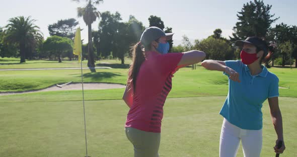 Two caucasian women playing golf wearing face masks greeting each other with elbows alt