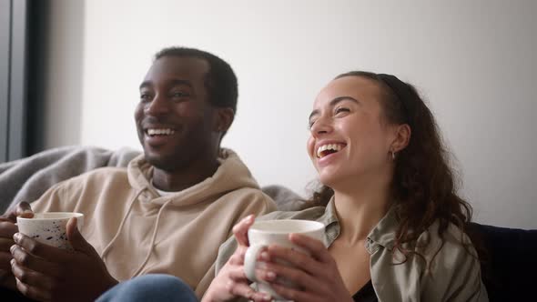 Smiling Young Couple With Hot Drinks Relaxing At Home Sitting On Sofa Watching On Demand TV Together