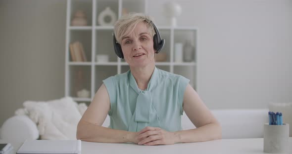 Charming Middle-aged Woman with Headphones Is Greeting By Videocall, Looking at Camera, Sitting in alt