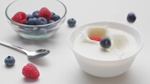 Raspberries and blueberry falling into white bowl with yogurt and whipped cream.