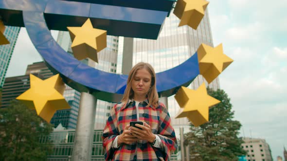 Lady Uses Mobile App on Smartphone Walking By Euro Sign in Business District