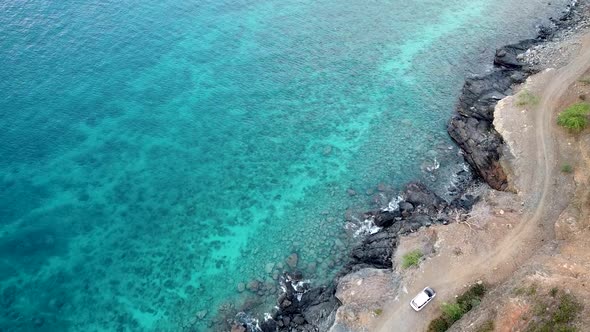 White car traveling slowly on a winding, rough and bumpy coastal road with beautiful turquoise water alt