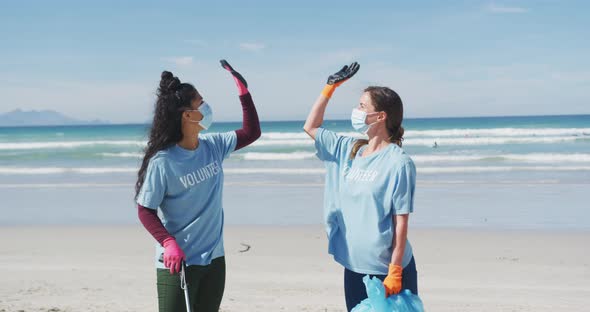 Two diverse women wearing volunteer t shirts and face masks picking up rubbish from beach alt