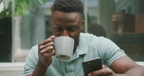 Happy african american man sitting with smartphone and drinking coffee in garden alt