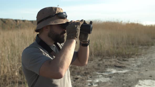 Outdoor Side Shot of Bearded Man Soldier in Hat and Military Tshirt Looking Through Binoculars alt