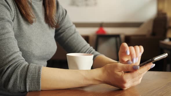 Woman Is Drinking a Cup of Coffee in Cafe and Typing a Message on Phone alt