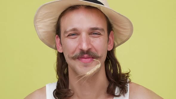 Long Darkhaired Happy Guy with Beautiful Smile in Beige Hat Taking Root in His Mouth Over Yellow alt