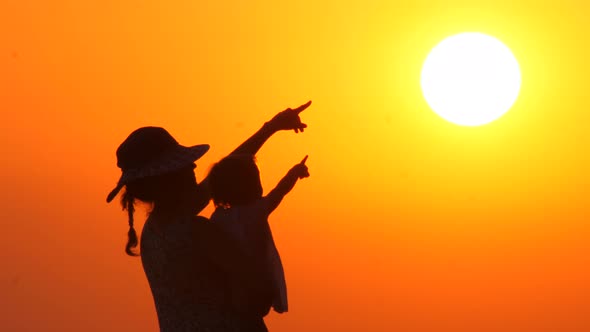 Mother in Hat with Child Looking and Pointing at the Sunset. Baby on Mothers Hands on Orange Sky