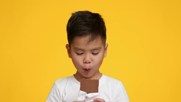Little Japanese Boy Eating Chocolate Bar Over Yellow Studio Background alt
