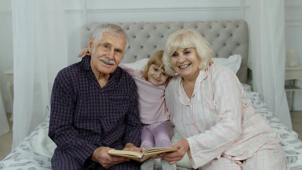 Cute Girl with Senior Retired Grandmother and Grandfather Sitting on Bed Reading Book in Bedroom alt