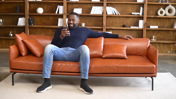 Excited Black Multiracial Man Sitting on the Brown Sofa Looking at the Phone alt