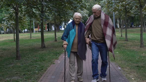 Mature Couple With Walking Sticks Walking Along Elderly Home Park, Support