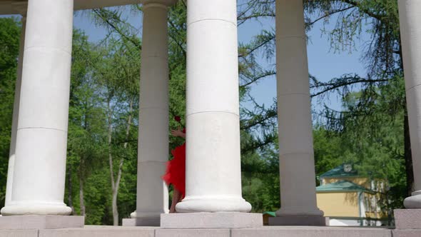 a Blonde with Big Breasts in a Red Fluffy Dress and with a Rose is Walking Through the Columns alt