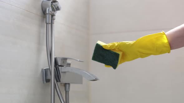 The hands of a woman in yellow gloves wash the faucet with a sponge and detergent.
